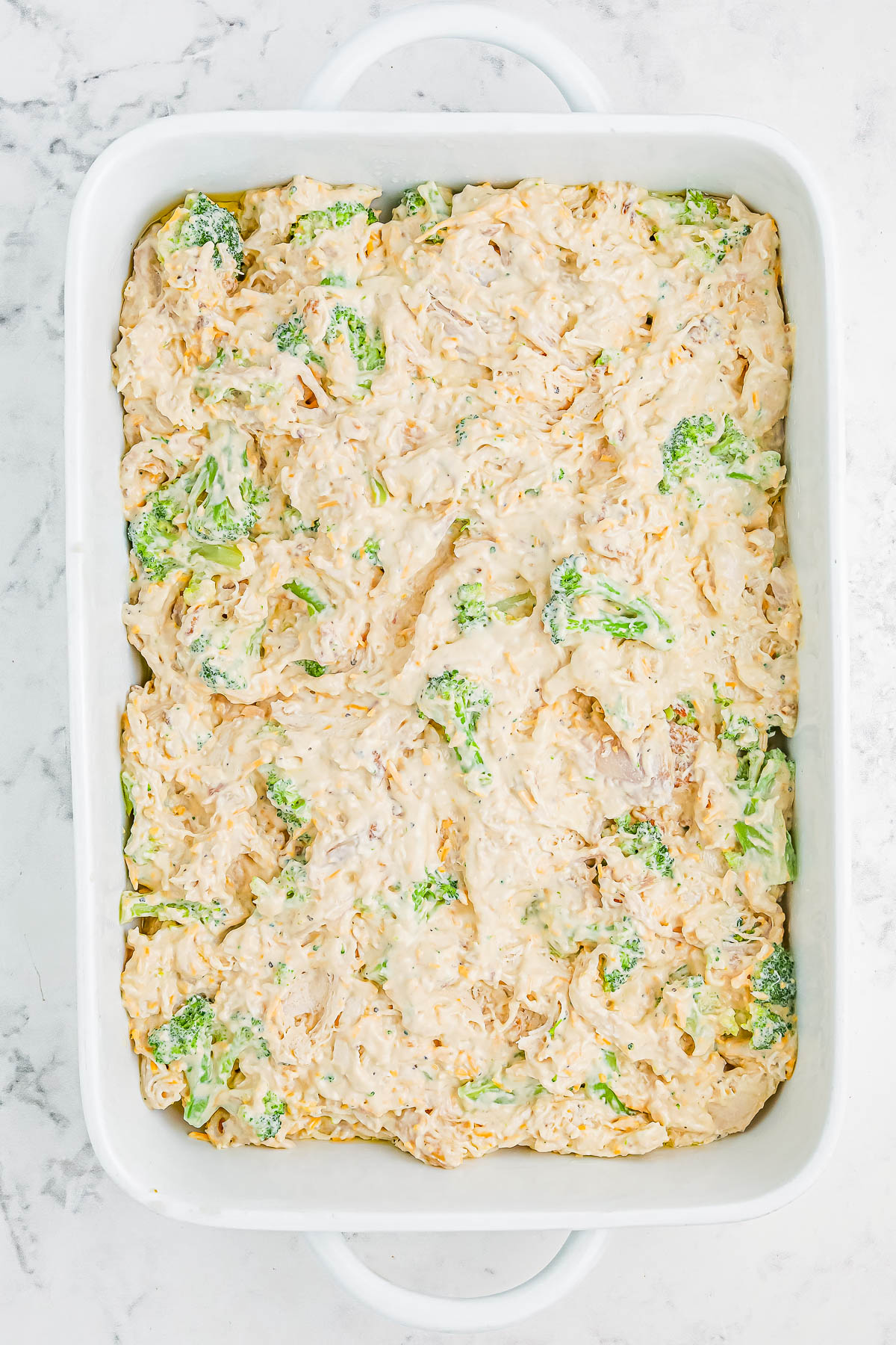 A white baking dish filled with a creamy casserole mixture, including visible pieces of broccoli, on a light marble surface.