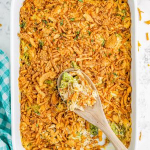 A casserole dish filled with a baked broccoli cheese casserole topped with crispy fried onions. A wooden spoon rests in the dish. A blue checkered cloth is on the side.