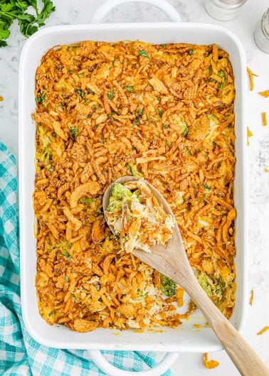 A casserole dish filled with a baked broccoli cheese casserole topped with crispy fried onions. A wooden spoon rests in the dish. A blue checkered cloth is on the side.