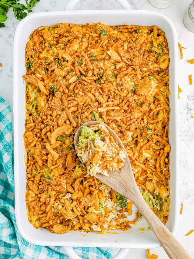 A casserole dish filled with a baked broccoli cheese casserole topped with crispy fried onions. A wooden spoon rests in the dish. A blue checkered cloth is on the side.