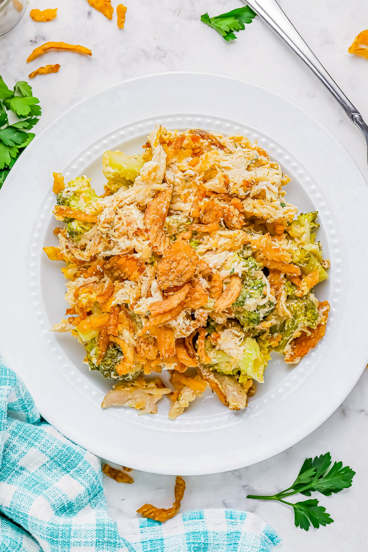 A white plate with a mixed salad of pasta, shredded chicken, and broccoli. Crushed chips are sprinkled on top. A blue checkered cloth is on the side, with parsley as garnish.