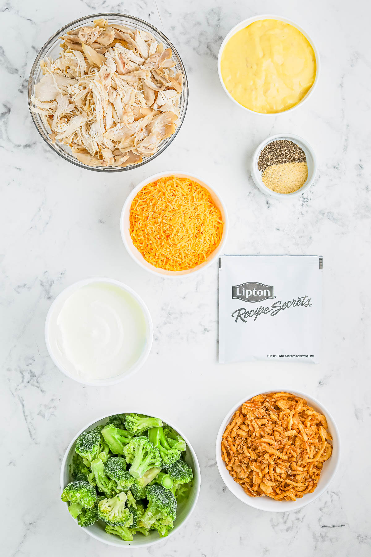 Ingredients on a marble surface: shredded chicken, condensed soup, spices, shredded cheese, sour cream, soup mix, broccoli, and crunchy topping.