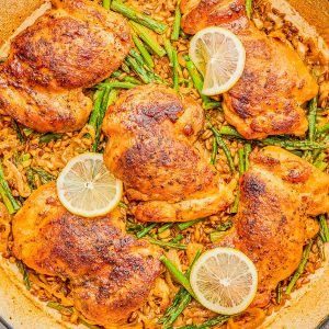 A large skillet with cooked chicken thighs, asparagus, lemon slices, and a rice dish, seasoned and arranged neatly.