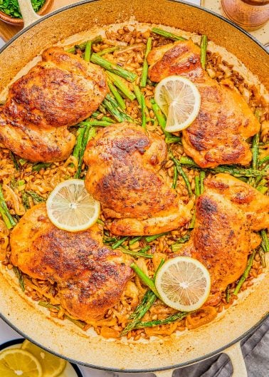 A large skillet with cooked chicken thighs, asparagus, lemon slices, and a rice dish, seasoned and arranged neatly.