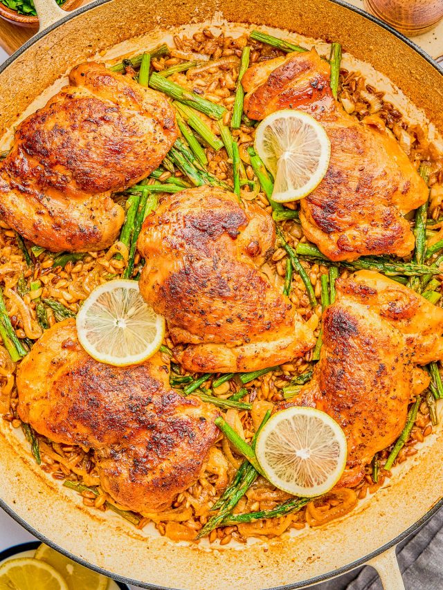 A large skillet with cooked chicken thighs, asparagus, lemon slices, and a rice dish, seasoned and arranged neatly.