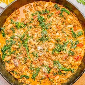 A skillet dish of ground meat, white beans, kale, and tomatoes in a creamy sauce, garnished with grated cheese, surrounded by lemon slices, red pepper flakes, and fresh herbs.