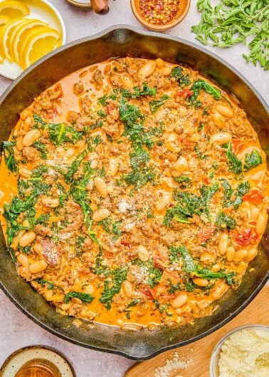 A skillet dish of ground meat, white beans, kale, and tomatoes in a creamy sauce, garnished with grated cheese, surrounded by lemon slices, red pepper flakes, and fresh herbs.