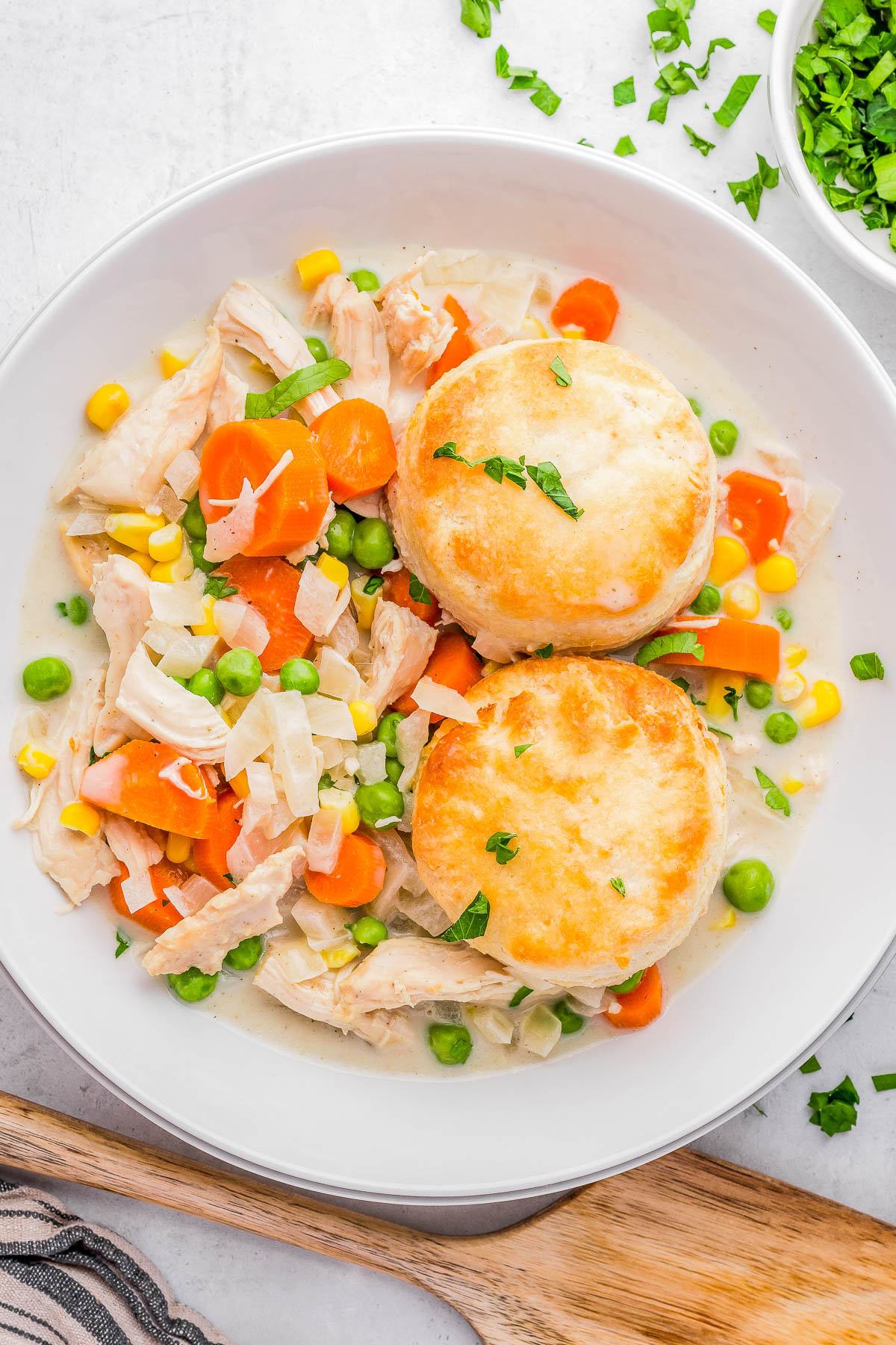A white plate contains a chicken and vegetable stew with carrots, peas, corn, and two round biscuits on top. Chopped herbs are sprinkled over the dish.