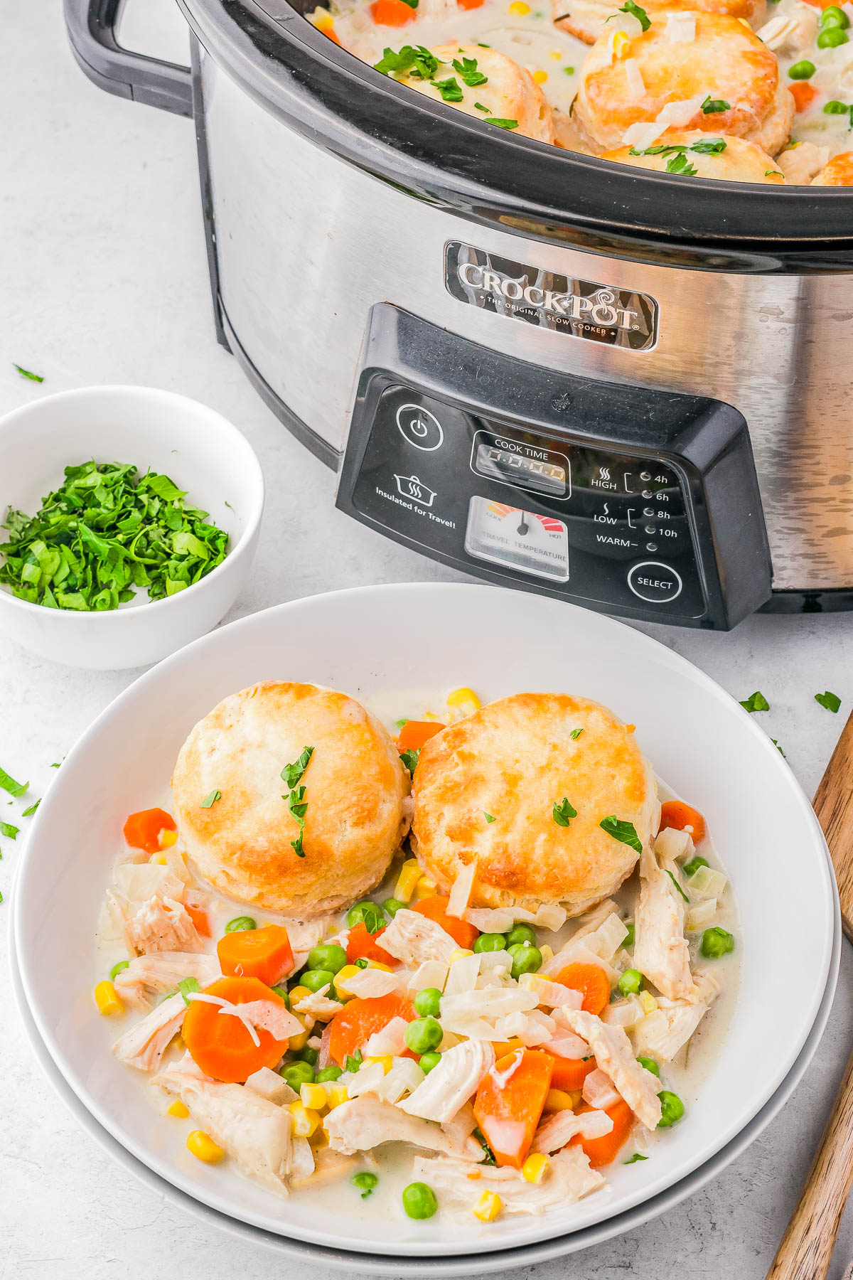 Slow cooker with chicken and vegetable stew topped with biscuits. A bowl of the stew with biscuits is in the foreground. A small bowl of chopped herbs is beside the cooker.