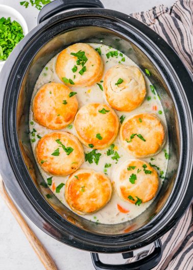 Slow cooker with creamy chicken stew topped with golden biscuits and garnished with chopped parsley.