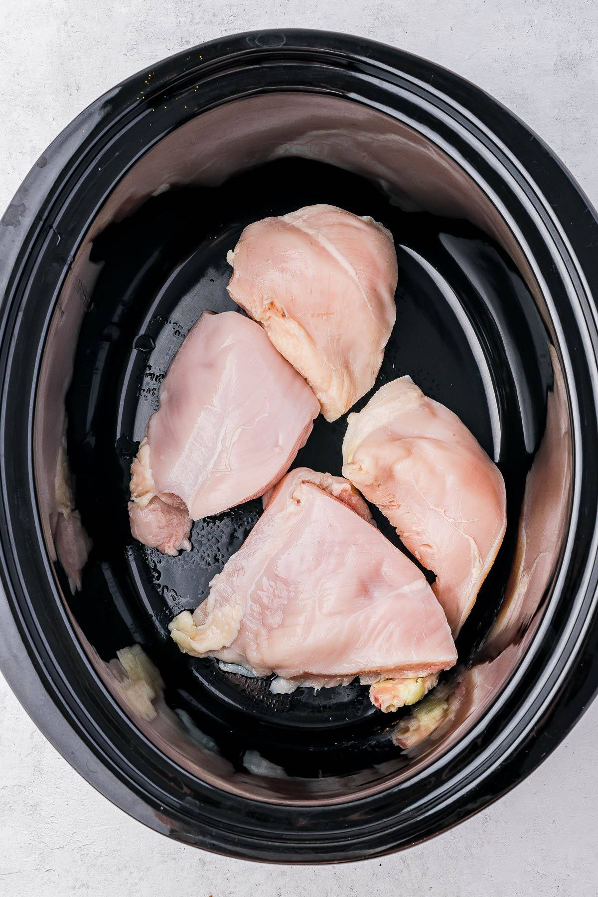 Four raw chicken breasts arranged in a slow cooker on a gray countertop.