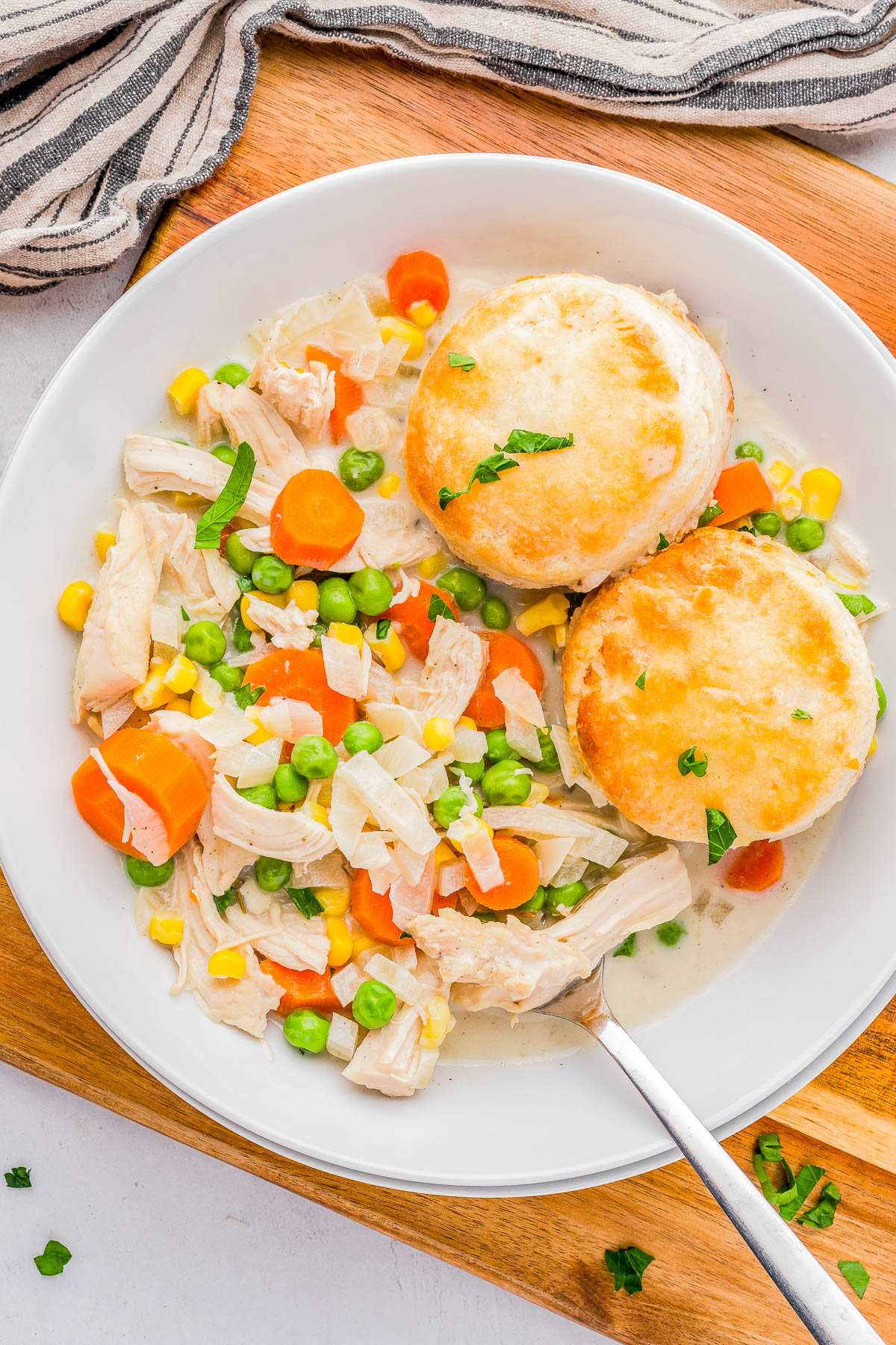 A plate of chicken pot pie filling with carrots, peas, corn, and flaky biscuits on a wooden board.