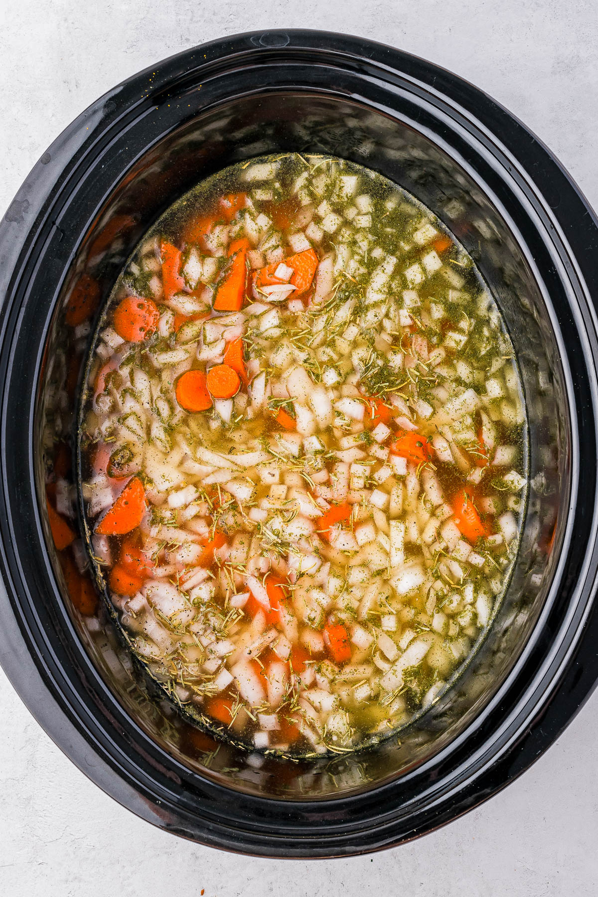 Slow cooker filled with uncooked soup ingredients, including chopped carrots, onions, herbs, and broth.
