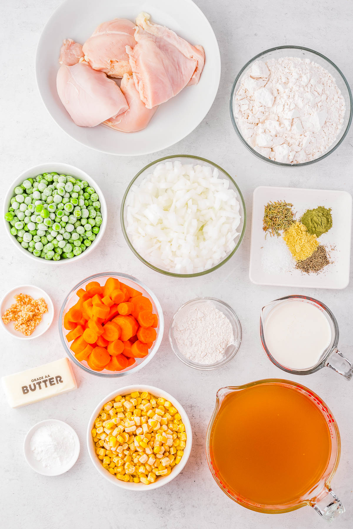 Ingredients for a chicken pot pie on a white surface, including raw chicken, flour, peas, onions, carrots, corn, butter, broth, milk, garlic, and assorted spices in bowls and a jug.