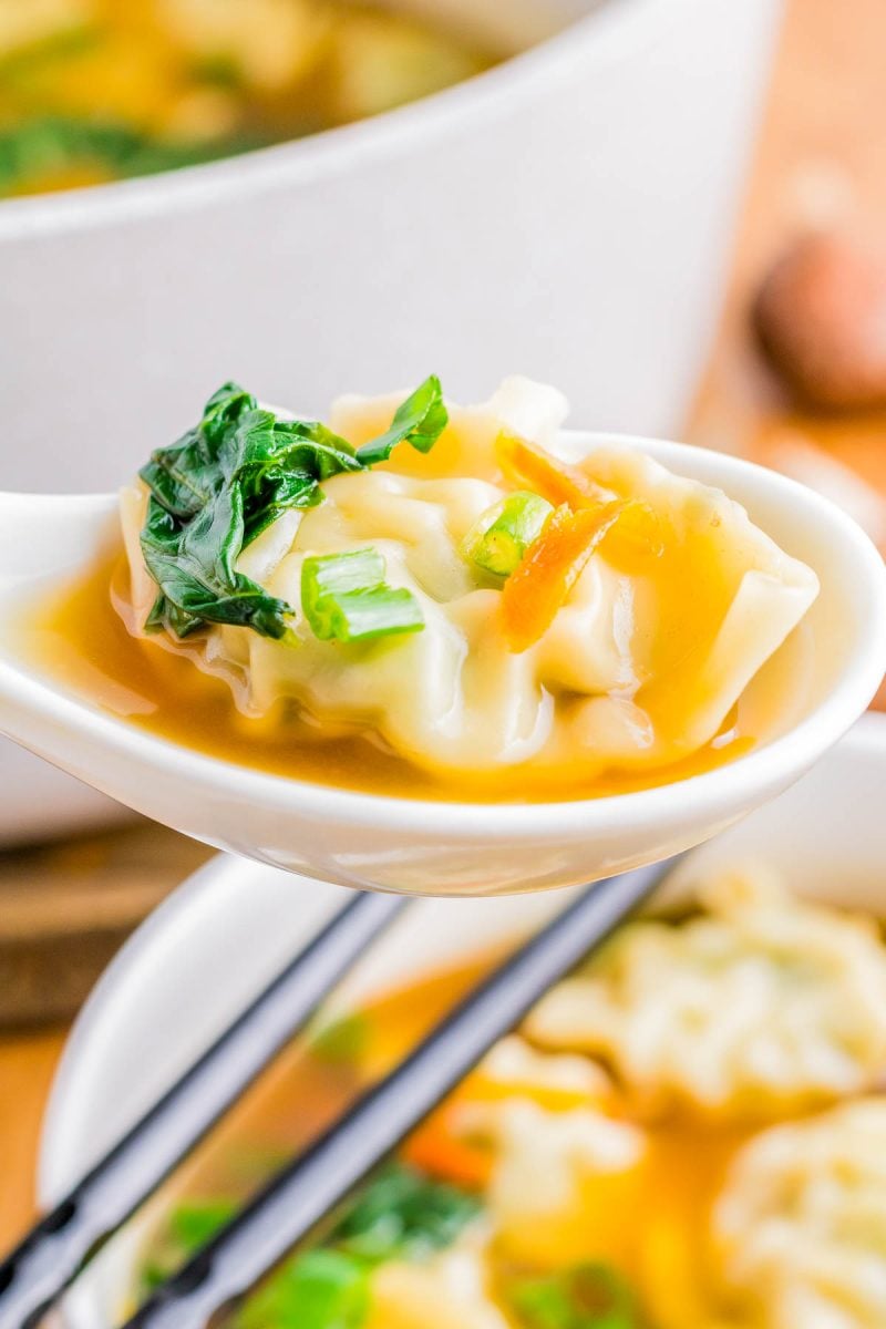 A spoon holding a dumpling in broth, garnished with greens, carrots, and a white bowl in the background.