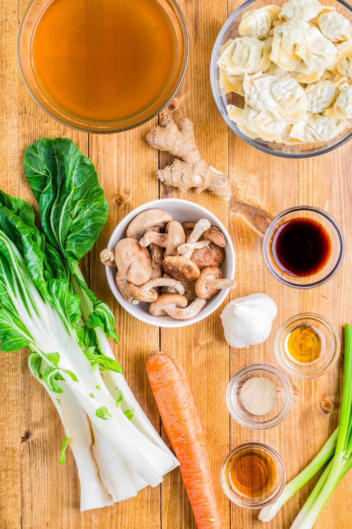 Ingredients for an Asian soup on a wooden surface: broth, dumplings, bok choy, mushrooms, ginger, garlic, soy sauce, a carrot, oil, and seasoning.