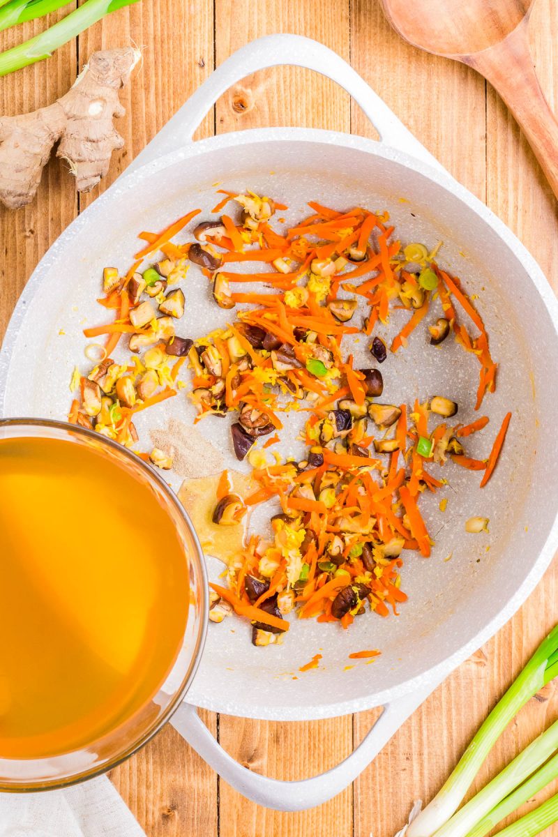 A skillet with sautéed carrots, mushrooms, and onions next to a bowl of broth on a wooden surface. Scallions and ginger root nearby.