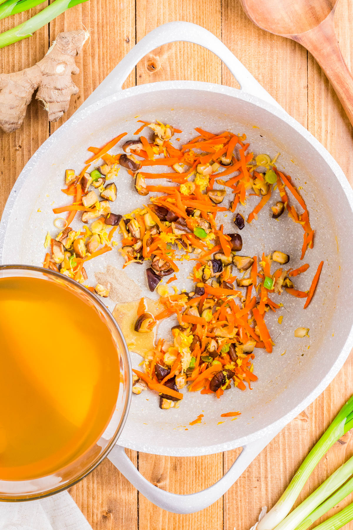 A skillet with sautéed carrots, mushrooms, and onions next to a bowl of broth on a wooden surface. Scallions and ginger root nearby.