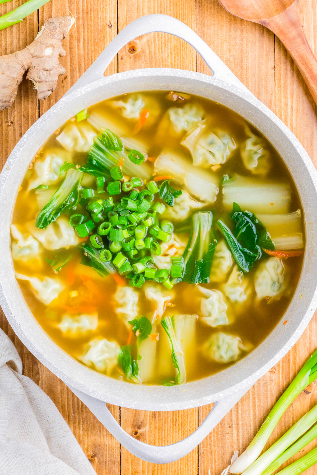 A pot of soup with dumplings, leafy greens, and chopped green onions on top, placed on a wooden table surrounded by fresh vegetables and a cloth napkin.