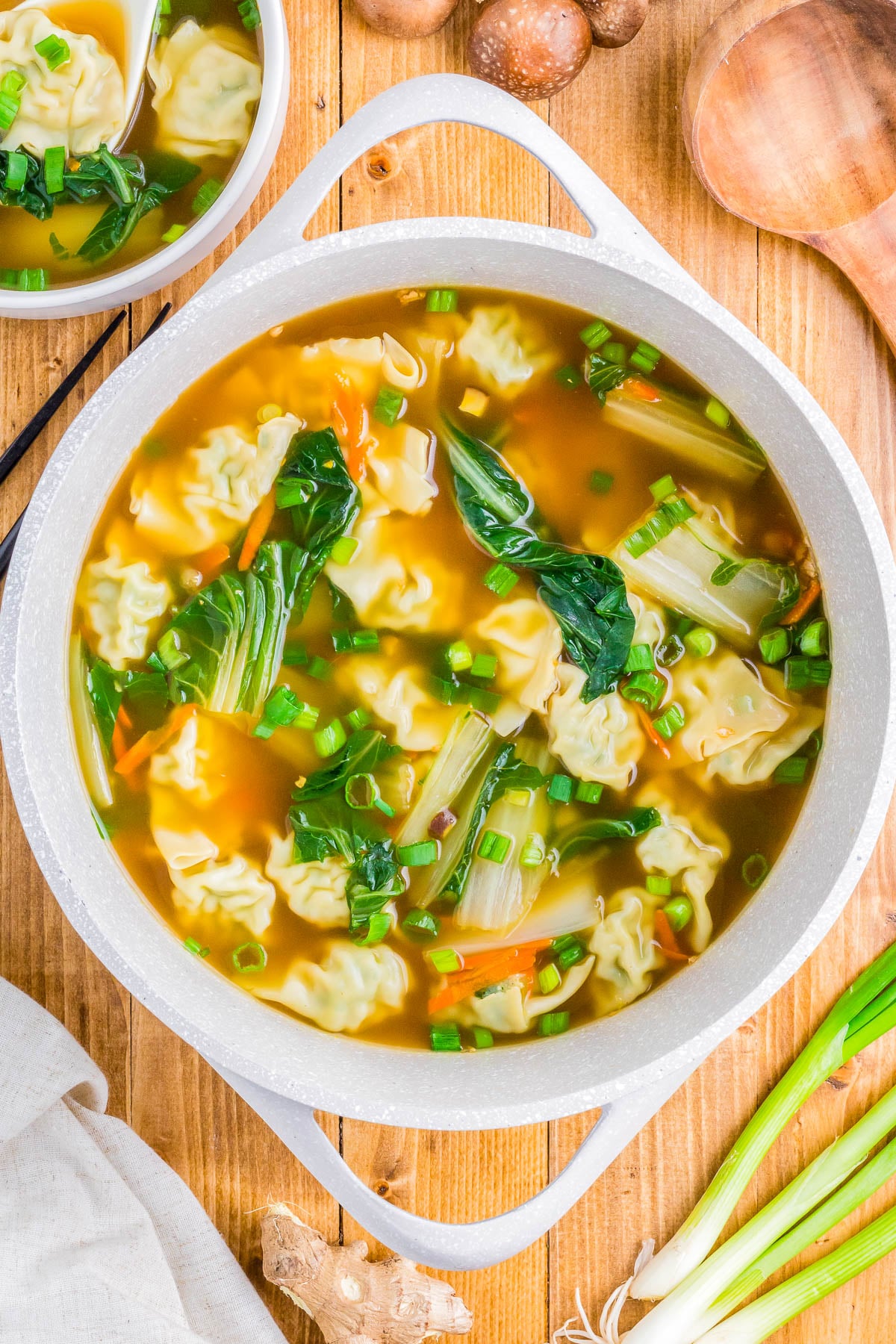 A pot of wonton soup with vegetables like bok choy and carrots, garnished with chopped green onions, placed on a wooden surface.