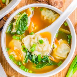 A bowl of soup with dumplings, spinach, and chopped green onions. A white spoon is lifting a dumpling. Shiitake mushrooms, ginger, and green onions surround the bowl.