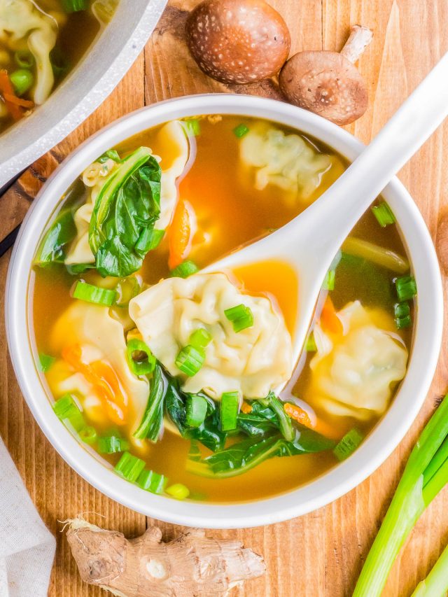 A bowl of soup with dumplings, spinach, and chopped green onions. A white spoon is lifting a dumpling. Shiitake mushrooms, ginger, and green onions surround the bowl.