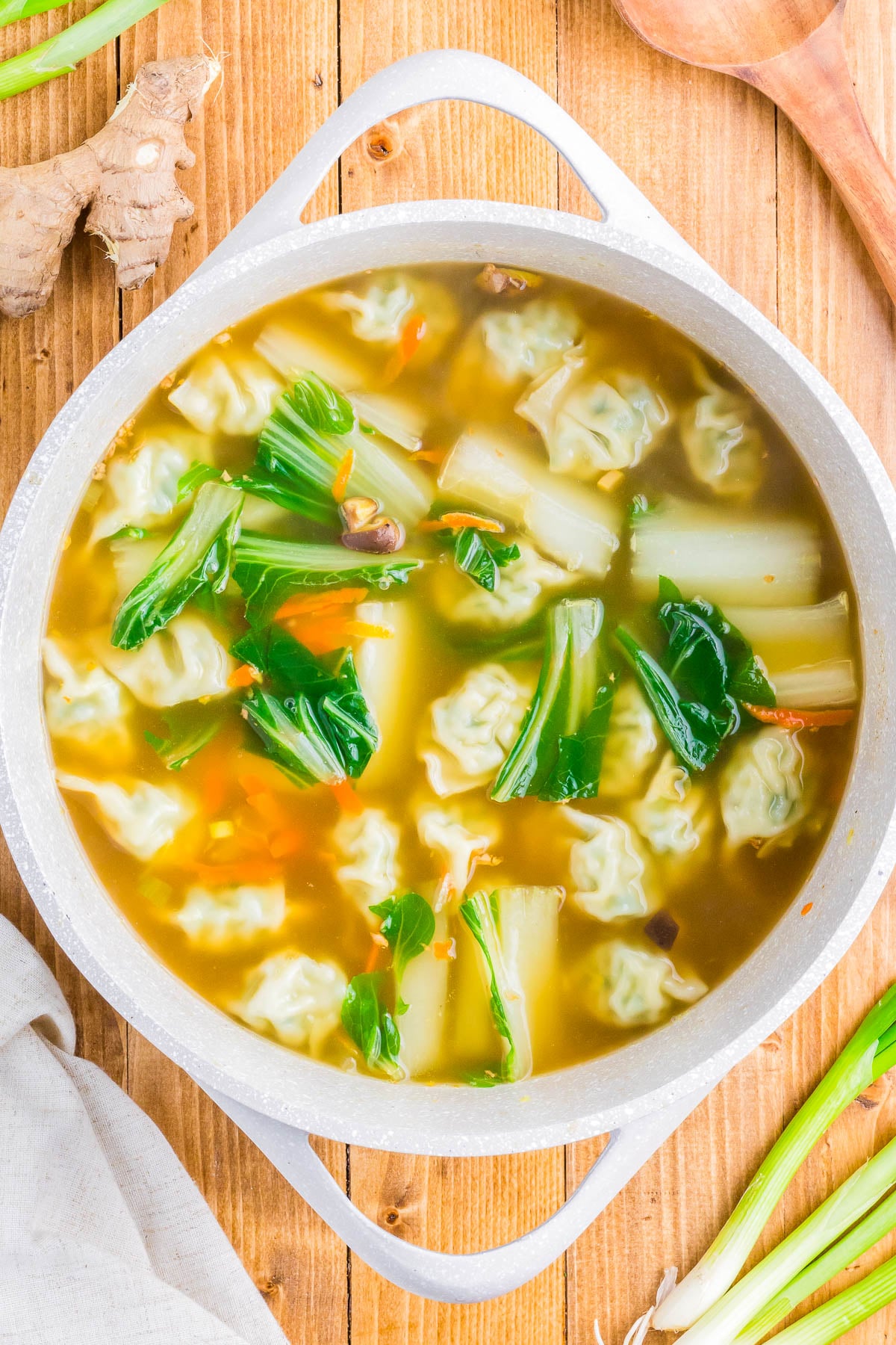A pot of soup with dumplings, bok choy, carrots, and broth on a wooden surface. Ginger and scallions are nearby.