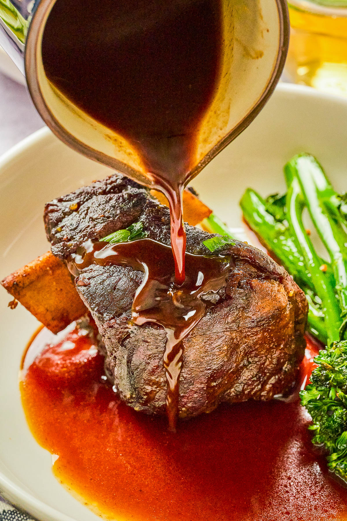 Beef short ribs with sauce being poured over, served with broccolini on a white plate.
