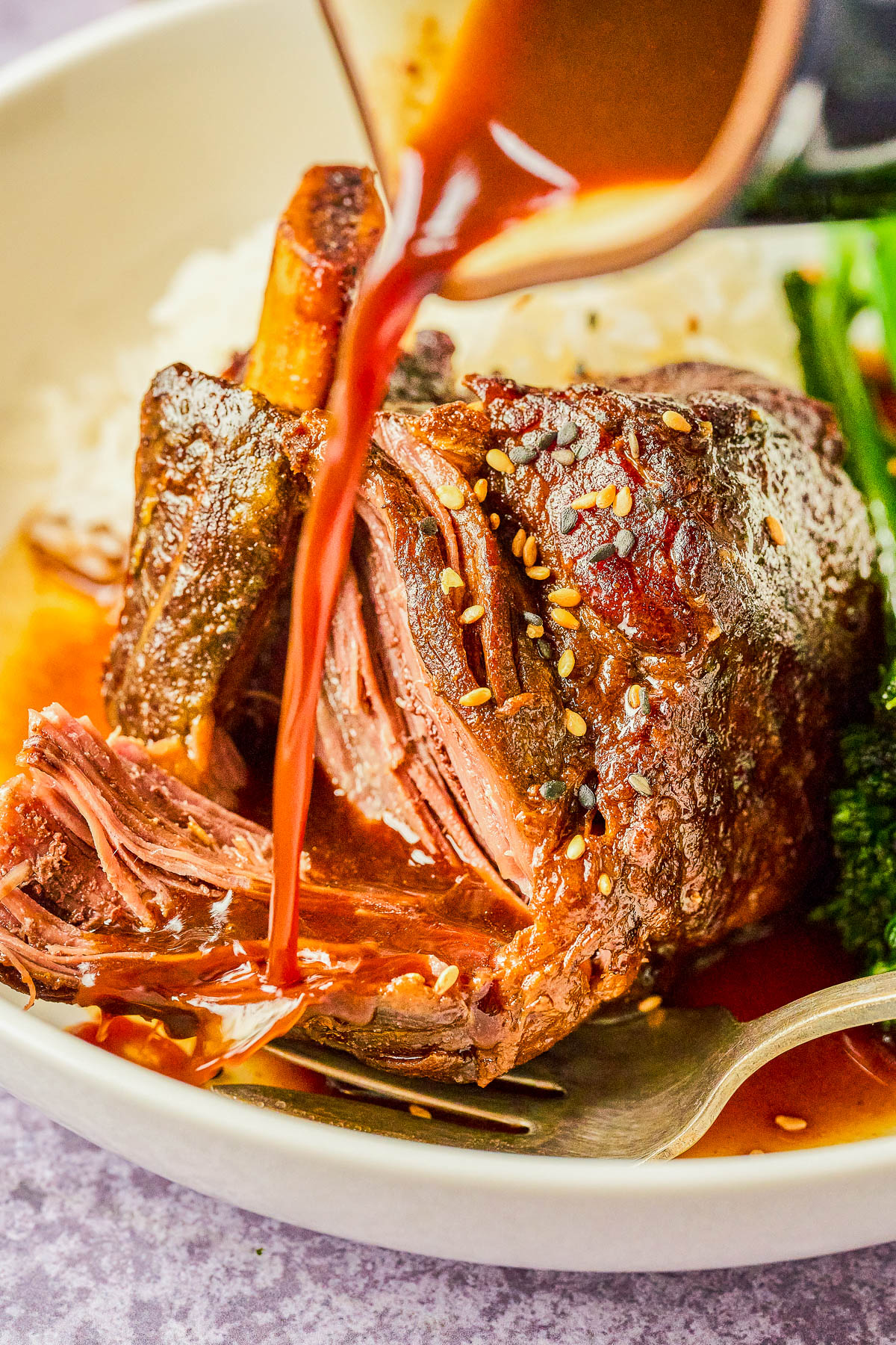 Sauce being poured over a braised beef shank served with rice and greens, garnished with sesame seeds, in a white bowl.