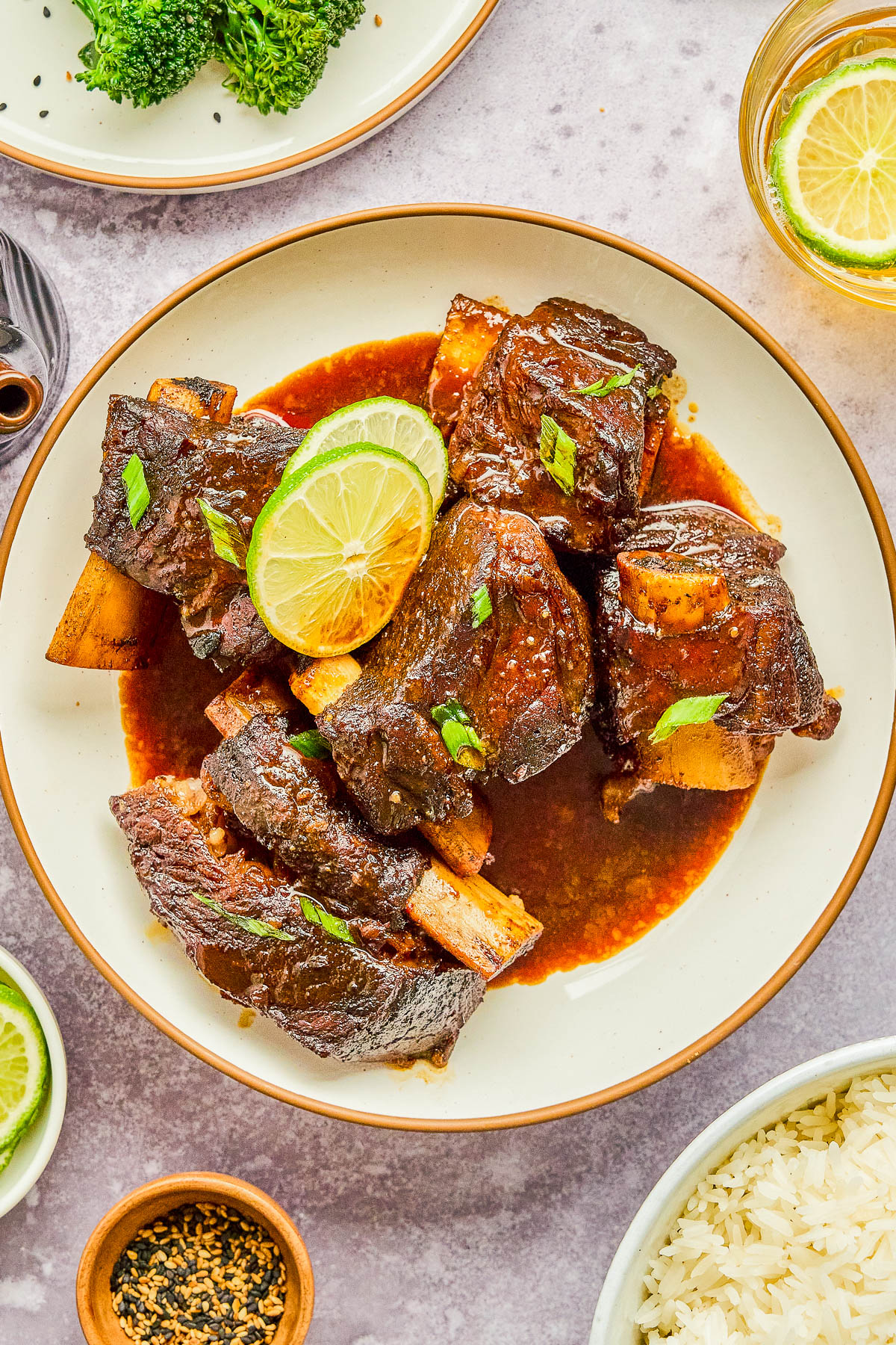 Plate of glazed beef ribs garnished with lime slices and herbs, surrounded by small dishes of rice, broccoli, spices, and a drink with a lemon slice.