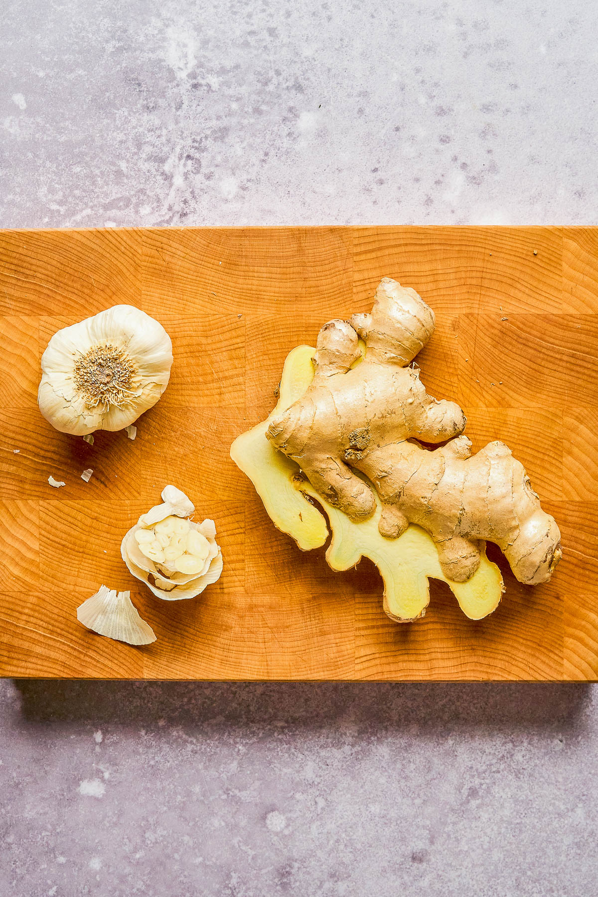 Ginger root with a slice cut off, and a whole garlic bulb with a few peeled cloves, on a wooden cutting board.