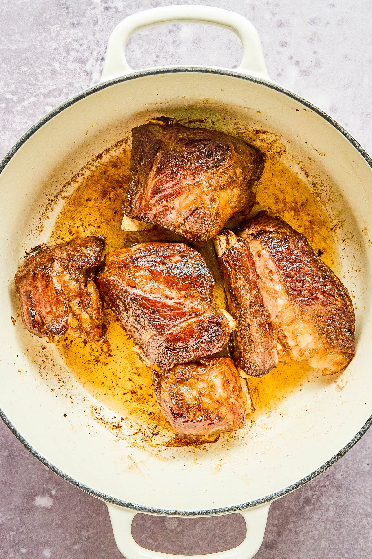 Chunks of browned meat cooking in a round, white pot on a light textured surface.