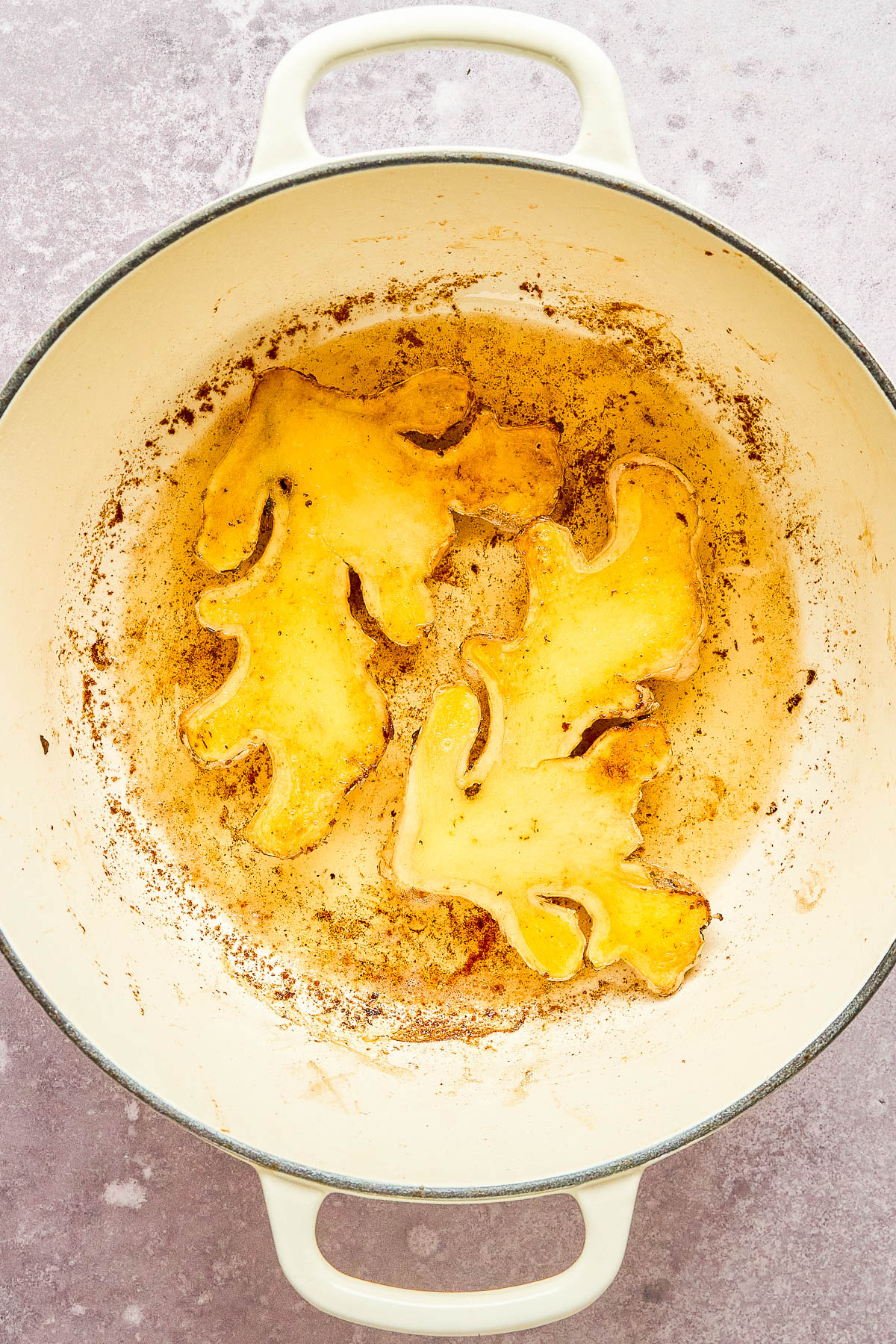 Two slices of ginger frying in a round, white ceramic pot. The ginger pieces are slightly browned, and the pot has some splatter marks. The surface and walls of the pot appear seasoned.