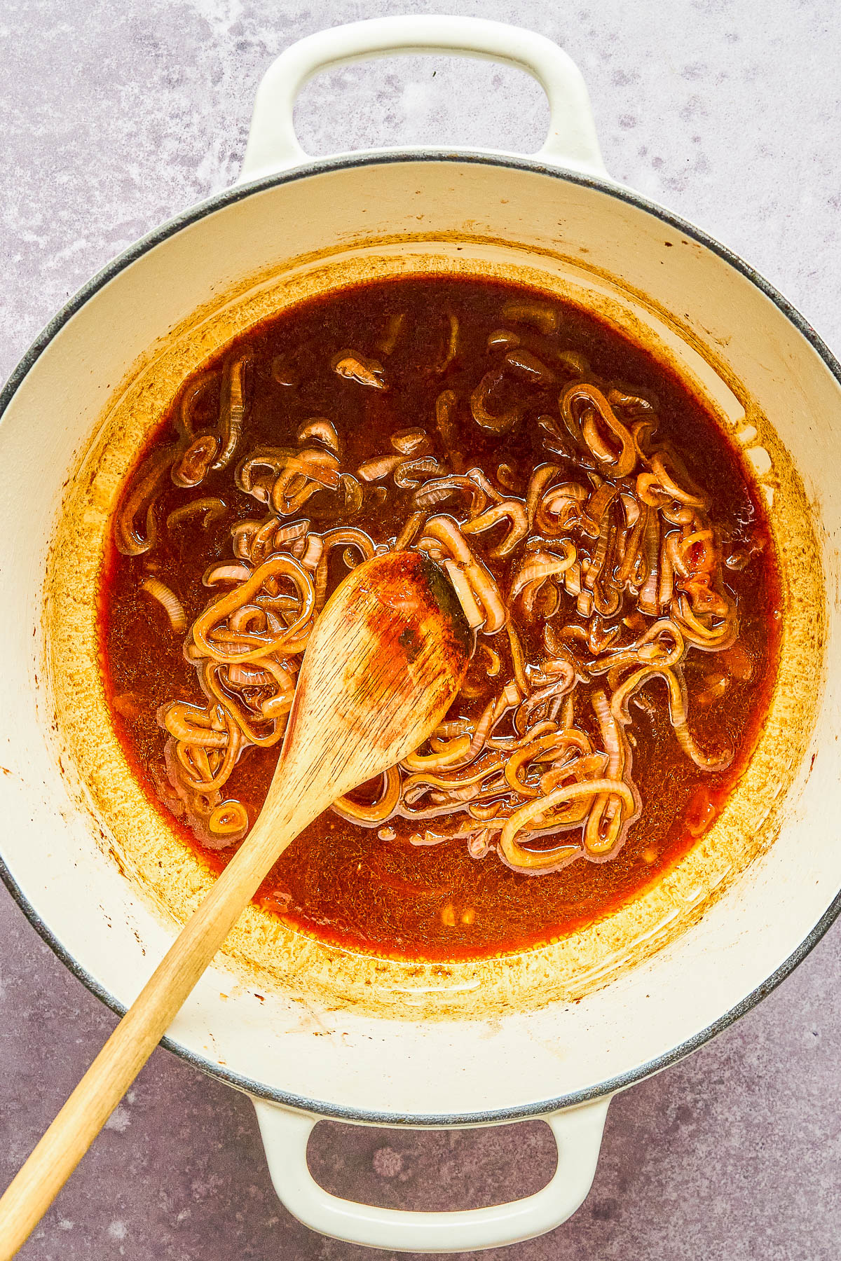 White pot with caramelized onions in a dark brown sauce, stirred with a wooden spoon, on a gray countertop.