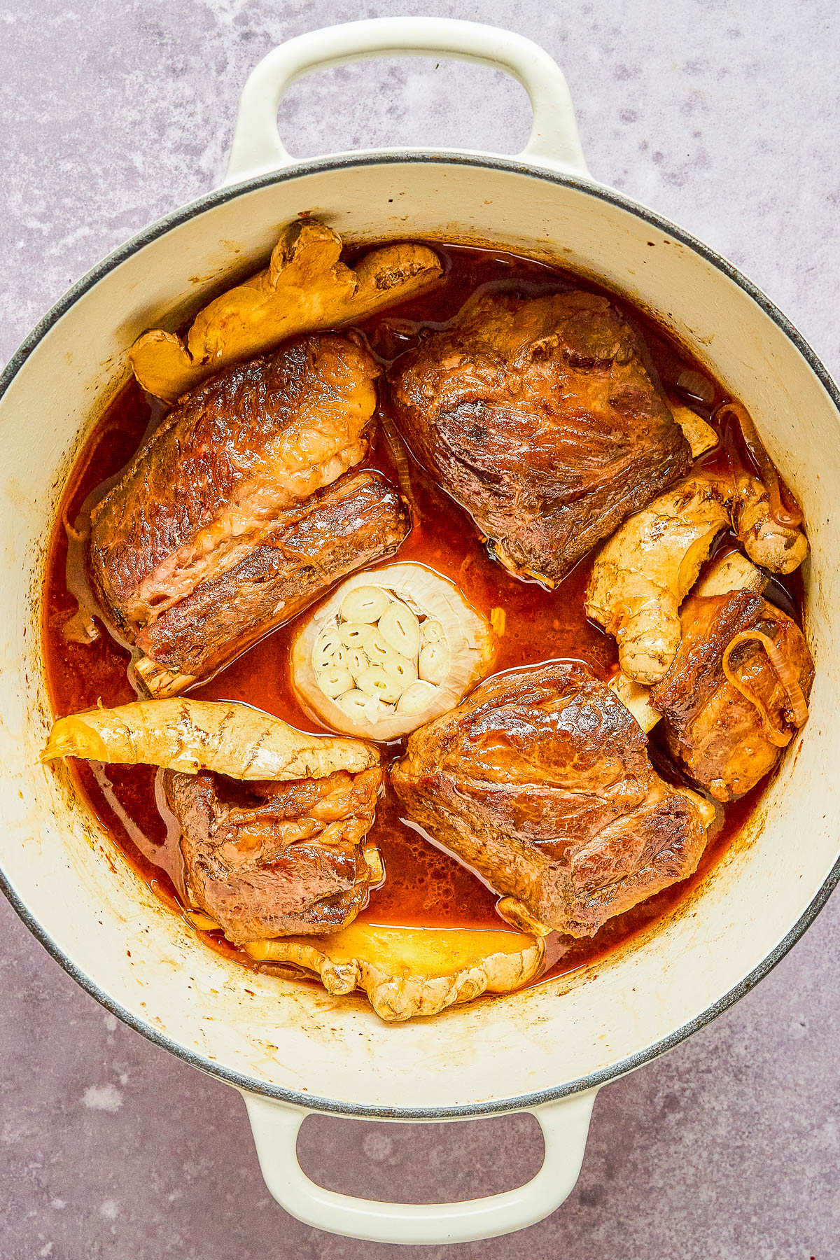 Pot of cooked beef with a whole garlic bulb and mushrooms in a red sauce.