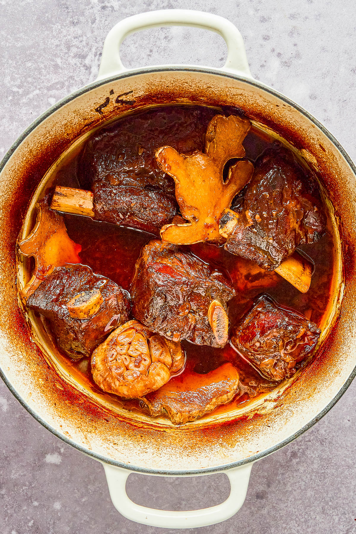 A pot filled with cooked beef short ribs, whole garlic, and mushrooms in a rich, dark sauce. The dish is in a white enamel pot on a gray textured surface.