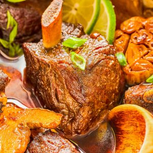 Close-up of braised beef short ribs with marrow bone in sauce, garnished with green onions and lime slices.