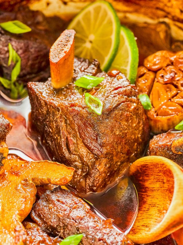 Close-up of braised beef short ribs with marrow bone in sauce, garnished with green onions and lime slices.