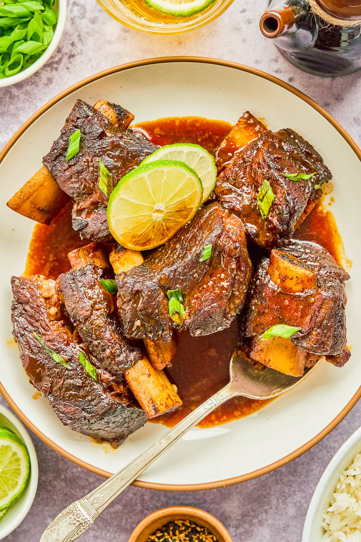 A plate of braised short ribs in sauce, garnished with lime slices and green onions. A silver spoon is placed on the plate. Sides of lime wedges, parsley, rice, and spices are visible.