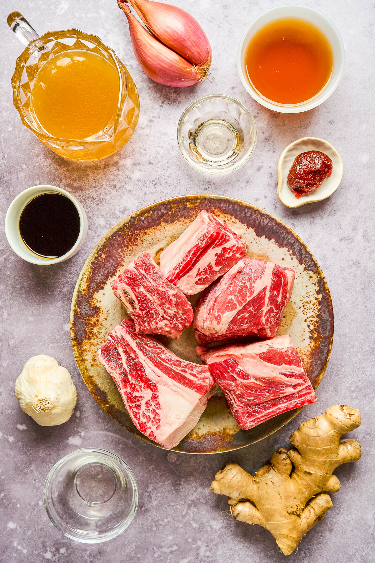 Raw beef short ribs on a plate surrounded by ingredients including garlic, ginger, soy sauce, broth, shallot, chili paste, honey, and oil on a white surface.