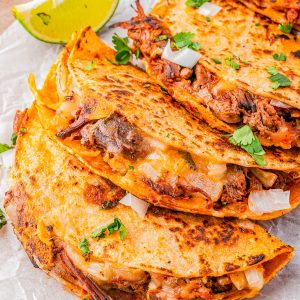 Three folded birria tacos filled with meat and melted cheese, garnished with chopped onions and cilantro, served with a lime wedge on parchment paper.