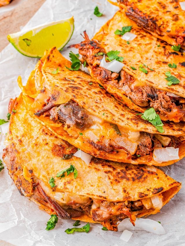 Three folded birria tacos filled with meat and melted cheese, garnished with chopped onions and cilantro, served with a lime wedge on parchment paper.