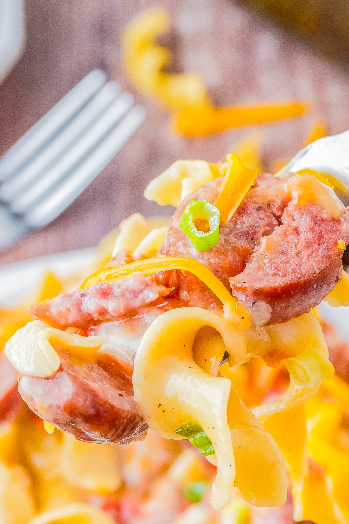A fork holding a bite of sausage, egg noodles, melted cheese, and sliced green onions, with a blurred plate and fork in the background.