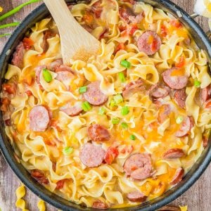 A skillet filled with creamy pasta, sliced sausage, tomatoes, and green onions, with a wooden spoon resting inside. Egg noodles are also visible next to the skillet.