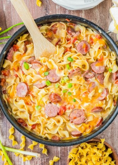 A skillet filled with creamy pasta, sliced sausage, tomatoes, and green onions, with a wooden spoon resting inside. Egg noodles are also visible next to the skillet.