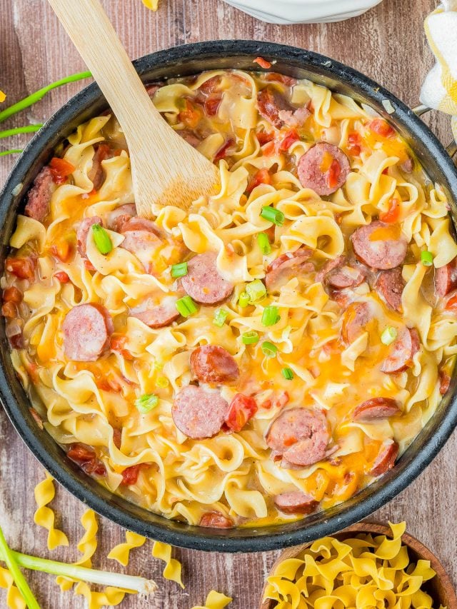 A skillet filled with creamy pasta, sliced sausage, tomatoes, and green onions, with a wooden spoon resting inside. Egg noodles are also visible next to the skillet.
