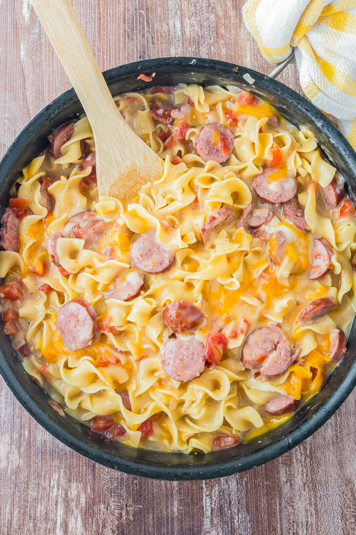 A skillet filled with egg noodles, sliced sausage, diced tomatoes, and bell peppers in a creamy sauce, with a wooden spoon resting inside.