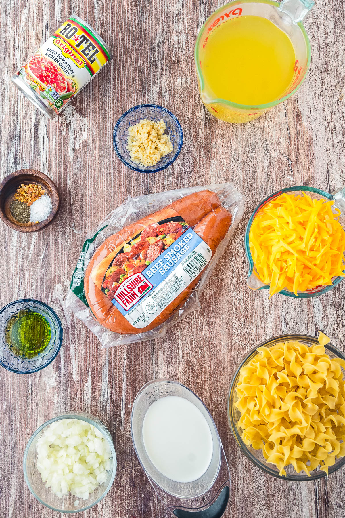 Ingredients for a beef smoked sausage pasta dish laid out on a wooden surface, including sausage, cheese, egg noodles, broth, milk, onion, garlic, oil, seasonings, and a can of tomatoes.
