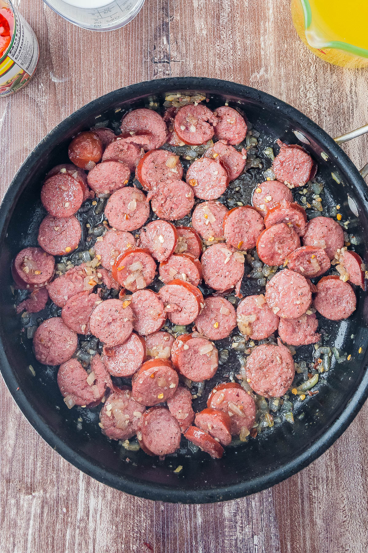 Sliced sausage cooking in a skillet with chopped onions and seasonings on a wooden surface.