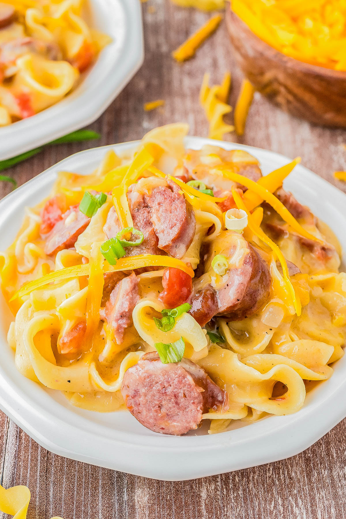 A plate of creamy pasta topped with sliced sausage, shredded cheese, diced tomatoes, and chopped green onions on a wooden table.